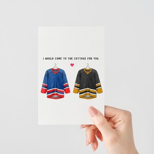 May include: A greeting card featuring illustrations of two hockey jerseys, one blue and red, the other black and gold, hanging on hangers. The text reads, "I would come to the cottage for you." A small pink heart is between the text and the jerseys.