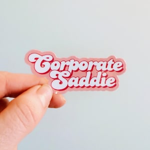May include: A pink sticker with the words "Corporate Saddie" in a retro font. The sticker has a white outline and is held by a hand against a light background. The text is in a bold, cursive style.