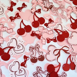 Op de afbeelding: Een verzameling uitgestanste papieren vormen met rode kersen, roze en wit geblokte strikken, hartvormige ringdoosjes met ringen erin en harten met een geschulpte rand. Het assortiment bevat diverse maten strikken en kersenparen in rood.