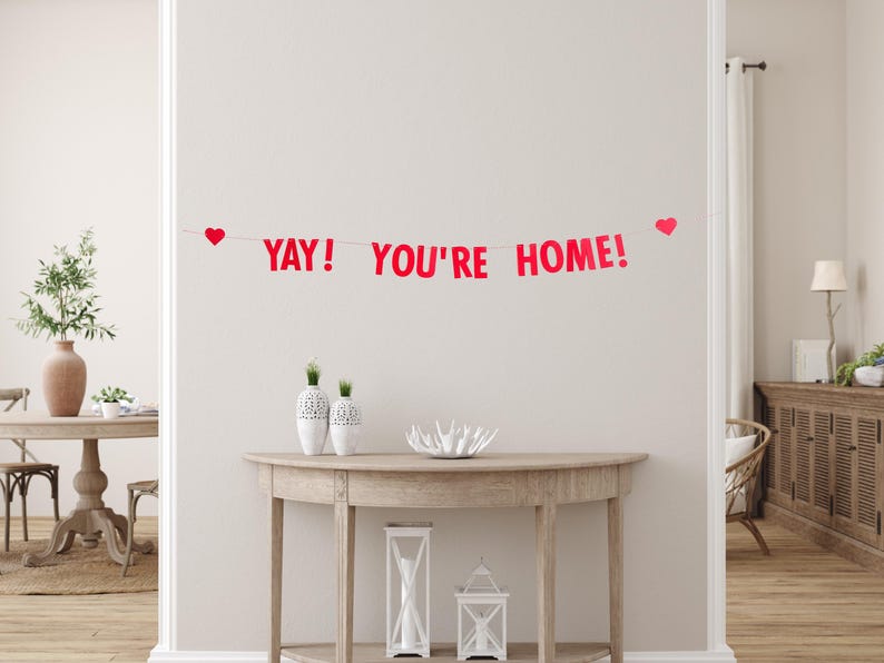 Pode incluir: Faixa de papel vermelha com o texto "YAY! YOU'RE HOME!" pendurada em uma parede branca. A faixa &eacute; decorada com cora&ccedil;&otilde;es vermelhos.
