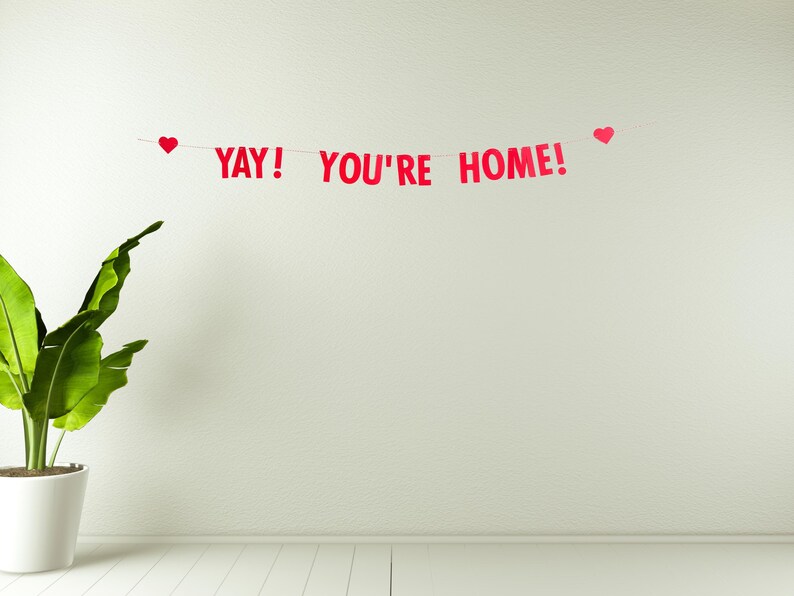 Pode incluir: Uma faixa vermelha com corda branca e cora&ccedil;&otilde;es vermelhos pendurados em uma parede branca. A faixa diz "YAY! YOU'RE HOME!"