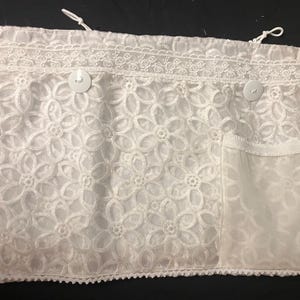 May include: White floral lace fabric with a scalloped edge and two white buttons. The fabric has a floral pattern and a pocket on the right side. Two white strings are at the top.