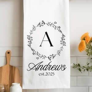 May include: White kitchen towel with a black floral wreath design, the letter "A" in the center, and the text "Andrews est. 2025" below.