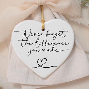 May include: White ceramic heart ornament with the text "Never forget the difference you make" in black cursive script. A small heart is drawn at the bottom of the ornament.