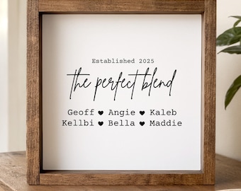 May include: A square wooden sign with a dark brown frame and a white center panel. The text reads "Established 2025", "the perfect blend", and lists the names "Geoff", "Angie", "Kaleb", "Kellbi", "Bella", and "Maddie" separated by black heart icons.