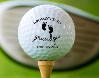 May include: A white golf ball with black text that reads "Promoted to grandpa February 2025" and a pair of footprints. The golf ball is on a tee and a golf club is in the background.