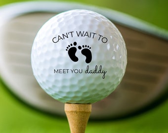May include: A white golf ball with black text that reads "Can't wait to meet you daddy" with a footprint symbol. The golf ball is on a tee and a golf club is behind it.
