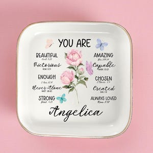 May include: A white ceramic trinket dish with a gold rim, featuring the words "YOU ARE" and inspirational words like "Beautiful" and "Amazing." Pink flowers and butterflies decorate the dish, with the name "Angelica" in script at the bottom.