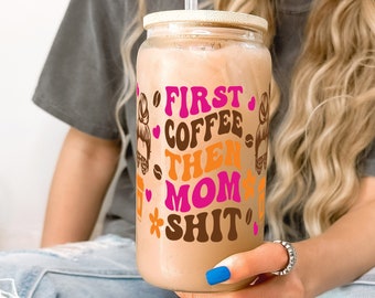 May include: A clear glass tumbler with a bamboo lid and a clear straw, filled with a coffee drink. The tumbler has the text "FIRST COFFEE THEN MOM SHIT" in pink, brown, and orange. The tumbler is held by a person wearing a grey t-shirt and blue jeans.