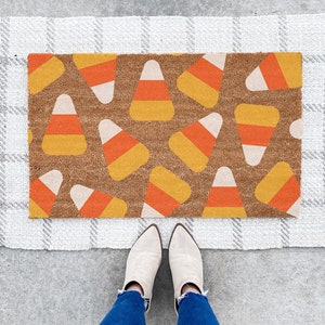 May include: A white doormat with a brown and orange candy corn pattern. The doormat has a fringe border and is on a grey concrete surface.