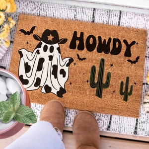 May include: A brown coir doormat with the text "Howdy" in black block letters. A black and white cow print ghost wearing a cowboy hat is on the left side of the mat. Two green cactus plants are on the right side of the mat.