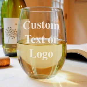 May include: A clear glass stemless wine glass with the text "Custom Text or Logo" etched on the side. The glass is filled with white wine and is sitting on a white surface.