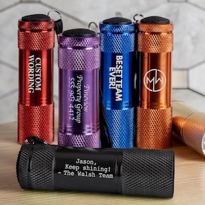 May include: A collection of personalized flashlights in various colors, including red, purple, blue, orange, and black. Each flashlight has custom text, such as "CUSTOM WORDING" and "BESET TEAM EVER!" One flashlight reads "Jason, Keep shining! - The Walsh Team."
