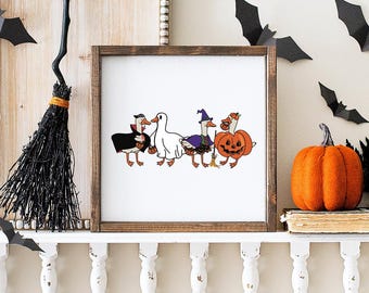 May include: A framed Halloween-themed print features four cartoon geese in costumes: a vampire, a ghost, a witch, and a jack-o'-lantern. The print is displayed on a white shelf with a black broom and paper bats. An orange pumpkin sits nearby.
