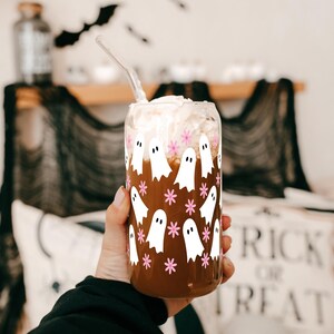 Hand holding a ghost-and-pink-flower glass cup filled with an iced drink near Halloween decorations.