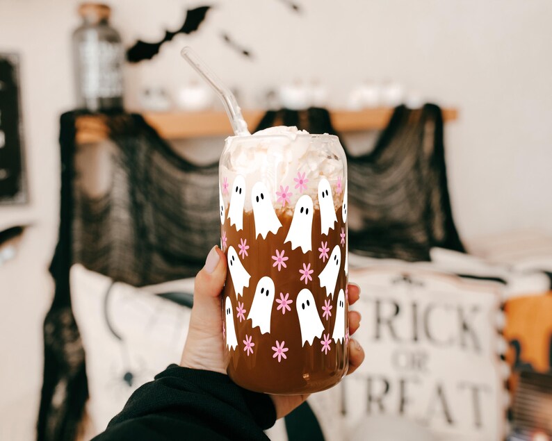 Hand holding a ghost-and-pink-flower glass cup filled with an iced drink near Halloween decorations.