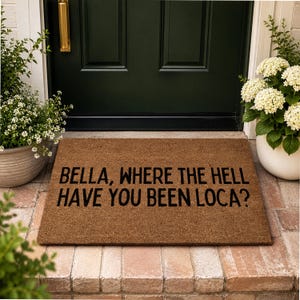 May include: A rectangular brown coir doormat featuring the black printed text BELLA, WHERE THE HELL HAVE YOU BEEN LOCA? The mat is positioned on a stone porch in front of a dark green door with a gold handle, flanked by two potted white hydrangea plants.