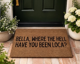 Puede incluir: Un felpudo rectangular de fibra de coco marrón con el texto impreso en negro BELLA, WHERE THE HELL HAVE YOU BEEN LOCA? La alfombra está situada en un porche de piedra frente a una puerta verde oscuro con pomo dorado, flanqueada por dos hortensias.
