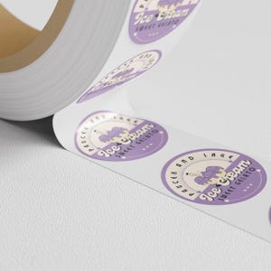 Close angled view of a roll of purple-and-cream circular stickers featuring sample logo artwork.