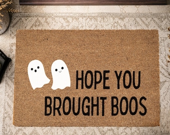 May include: A rectangular brown coir doormat featuring two white ghost illustrations on the left side. The text HOPE YOU BROUGHT BOOS is printed in bold black capital letters across the right side of the mat surface.