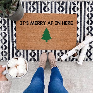 Overhead view of an It's merry AF in here doormat with a green tree, winter boots and a mug nearby.