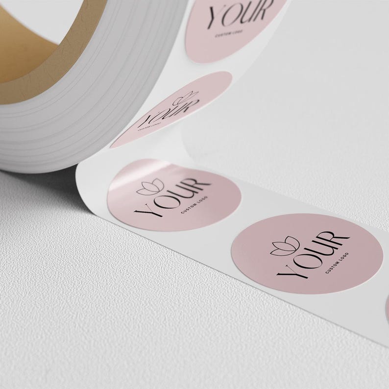Roll of circular pale pink stickers with a flower emblem and ‘Your custom logo,’ with several labels visible along the backing paper.
