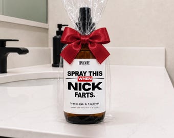 Puede incluir: Un frasco pulverizador de vidrio ámbar envuelto en plástico transparente con un lazo de satén rojo oscuro. La etiqueta dice: UNIQUE, SPRAY THIS WHEN NICK FARTS. Scent: Oak & Teakwood. El frasco contiene 236 ml y tiene un cabezal negro.