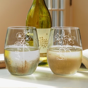 May include: Two clear glass wine glasses with white floral designs and names "Ashley" and "Kaitlin" etched on them. The glasses are filled with white wine and are sitting on a white surface.