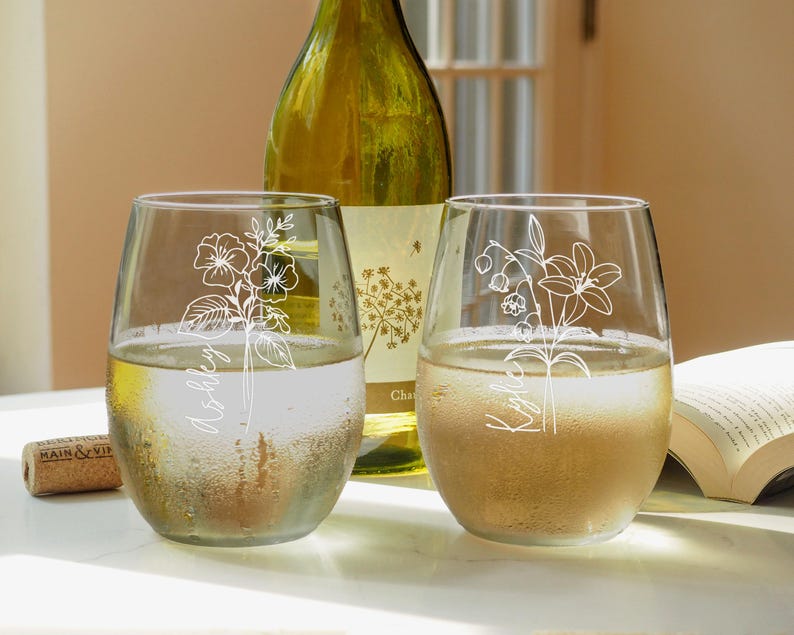 May include: Two clear glass wine glasses with white floral designs and names "Ashley" and "Kaitlin" etched on them. The glasses are filled with white wine and are sitting on a white surface.