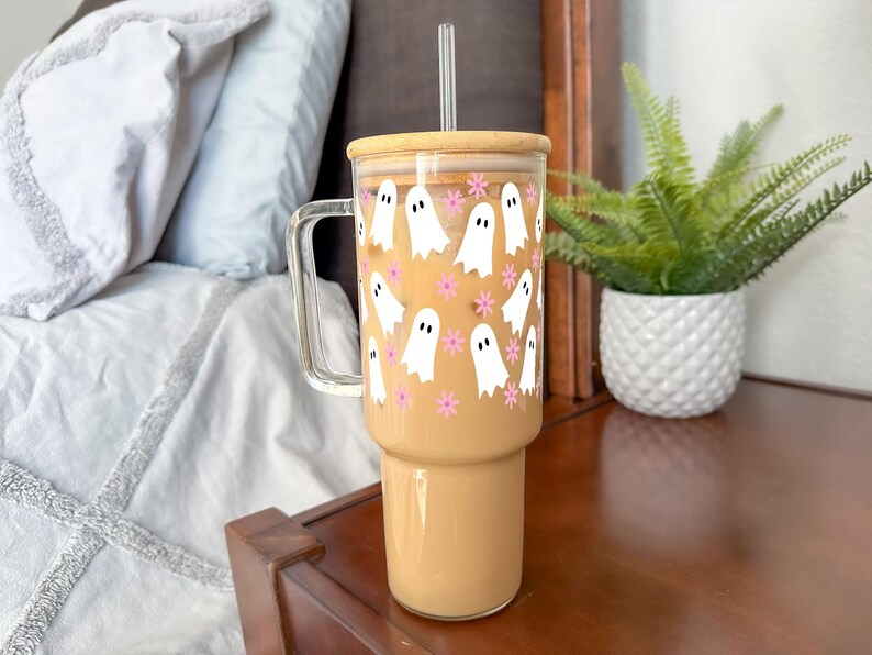 Large handled glass tumbler with white ghosts and pink flowers, filled with iced coffee on a bedside table.