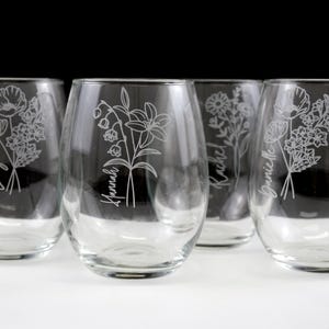 May include: Four clear glass wine glasses with etched floral designs and names, including "Lizzy", "Hannah", "Rachel", and "Jamie", on a white surface.