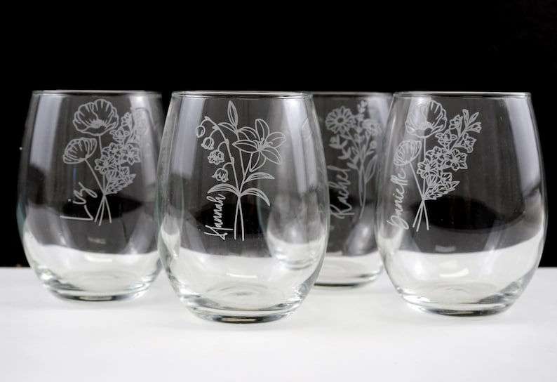 May include: Four clear glass wine glasses with etched floral designs and names, including "Lizzy", "Hannah", "Rachel", and "Jamie", on a white surface.