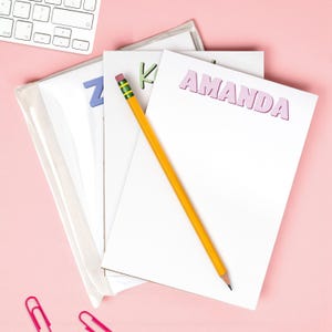 May include: A stack of white notepads with the name "AMANDA" in pink letters. A yellow pencil with a pink eraser rests on the notepad. The background is a light pink surface. A keyboard and paperclips are also visible.