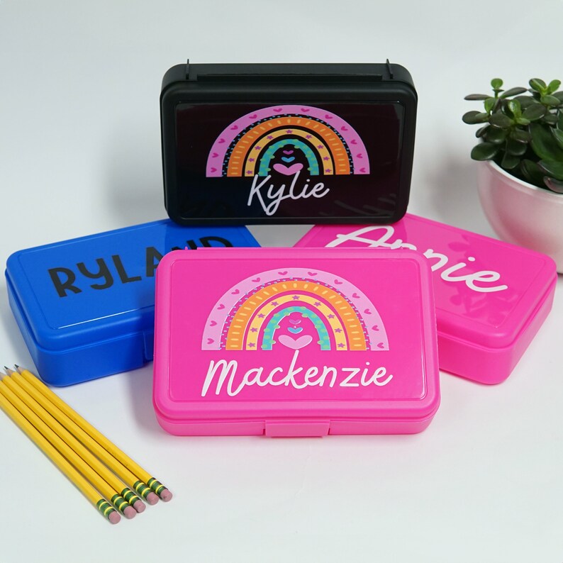 May include: Three colorful plastic pencil cases with rainbow designs and personalized names. The cases are blue, pink, and black. The names are Ryland, Mackenzie, and Kylie.