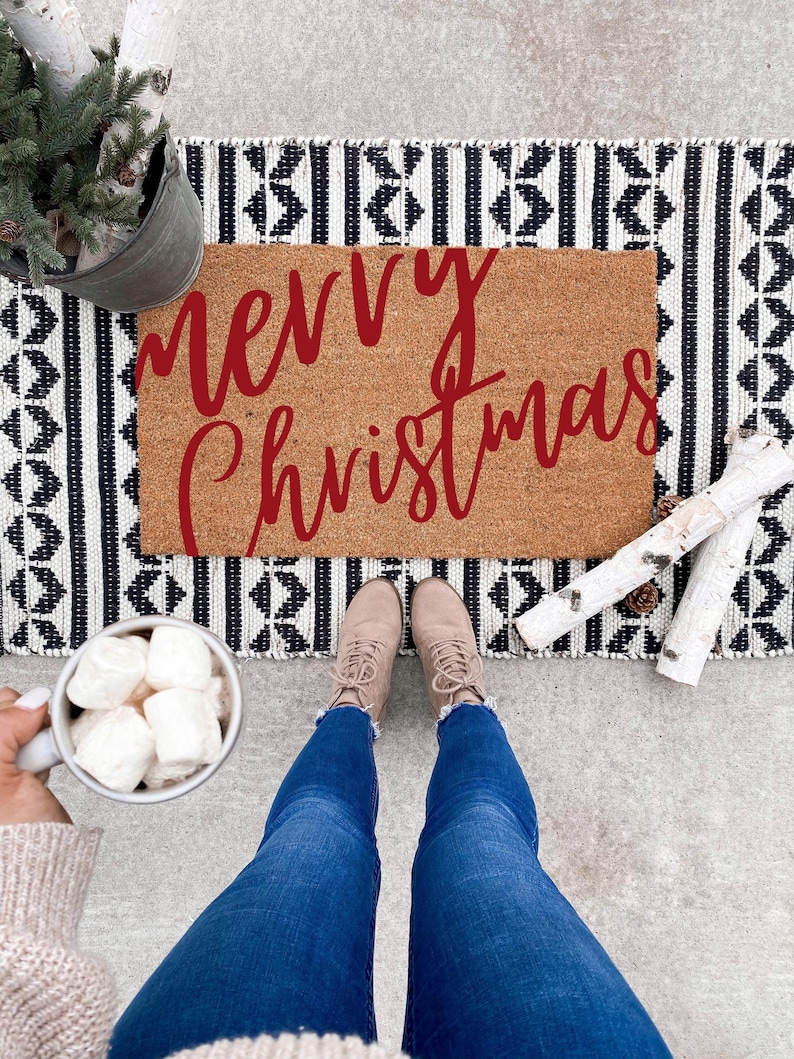 Overhead view of a red script Merry Christmas doormat with winter boots and a mug visible.