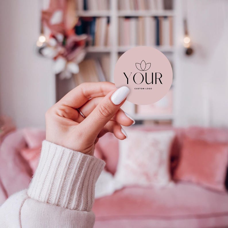 Hand holding a round pale pink sticker with a flower emblem and ‘Your custom logo,’ against a softly blurred pink studio background.