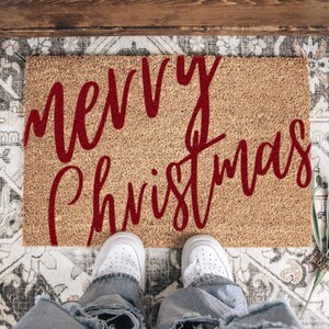 Red script Merry Christmas doormat viewed from above with white shoes at the edge.