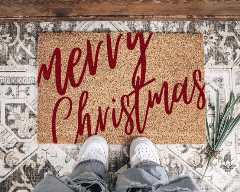 Red script Merry Christmas doormat viewed from above with white shoes at the edge.