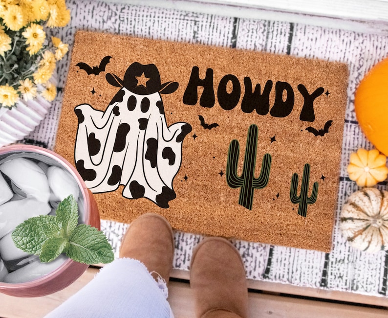 May include: A brown coir doormat with the text "Howdy" in black block letters. A black and white cow print ghost wearing a cowboy hat is on the left side of the mat. Two green cactus plants are on the right side of the mat.