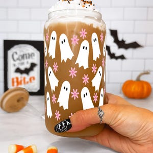 Hand holding a whipped-cream-topped drink in a ghost-and-flower glass cup beside candy corn and a pumpkin.
