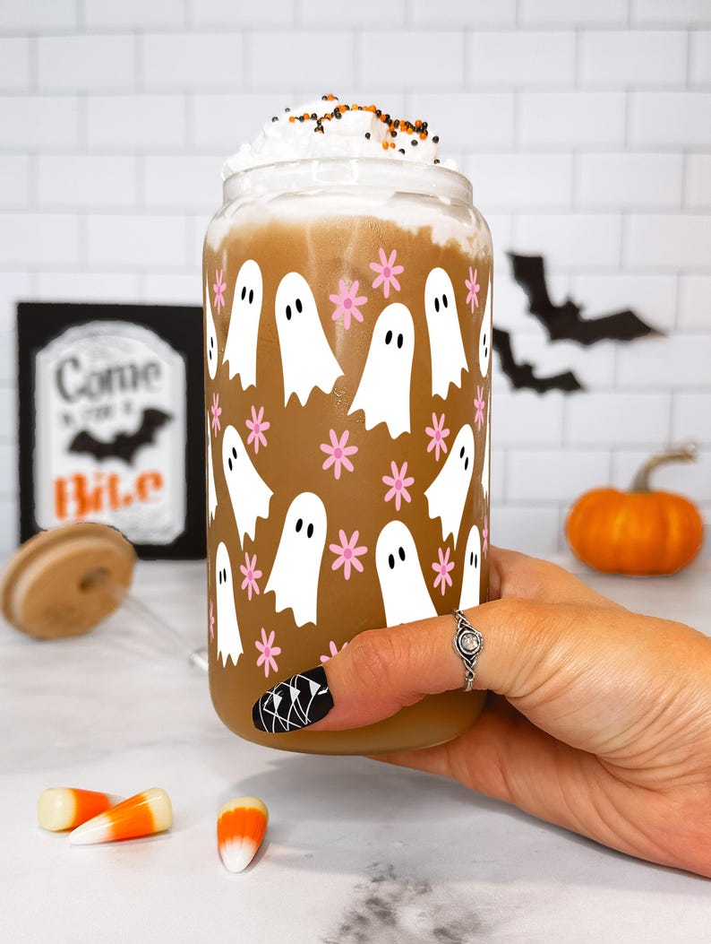 Hand holding a whipped-cream-topped drink in a ghost-and-flower glass cup beside candy corn and a pumpkin.