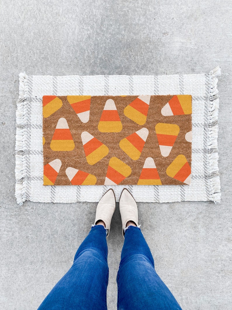 May include: A white doormat with a brown and orange candy corn pattern. The doormat has a fringe border and is on a grey concrete surface.