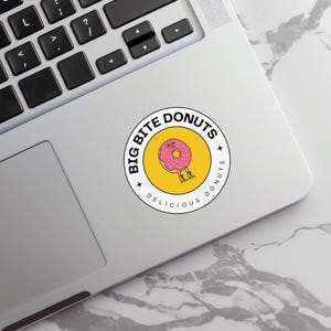 Round ‘Big Bite Donuts’ sticker with a pink doughnut illustration attached beside a laptop keyboard.