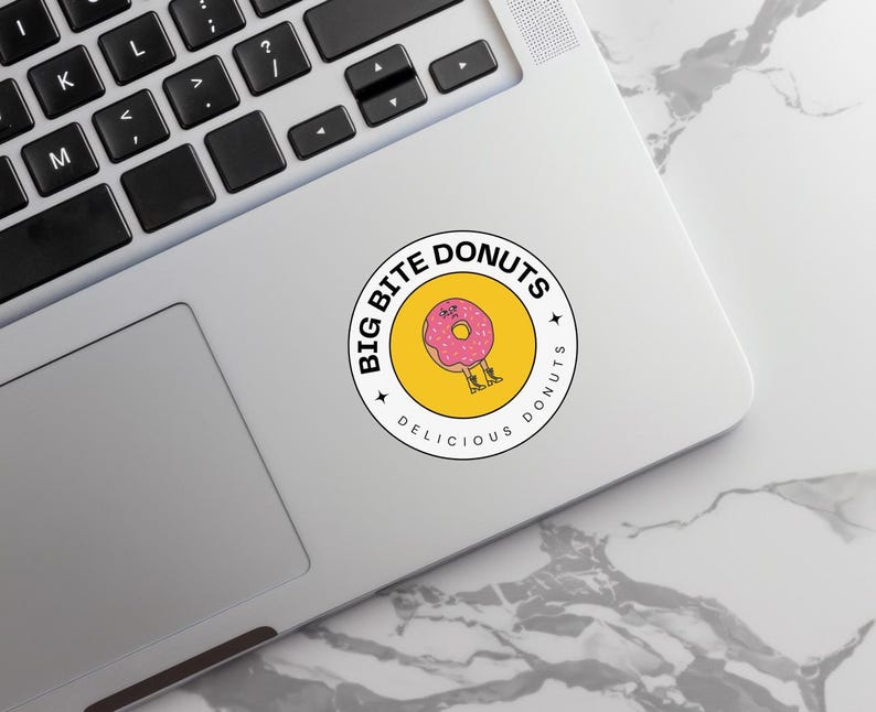 Round ‘Big Bite Donuts’ sticker with a pink doughnut illustration attached beside a laptop keyboard.