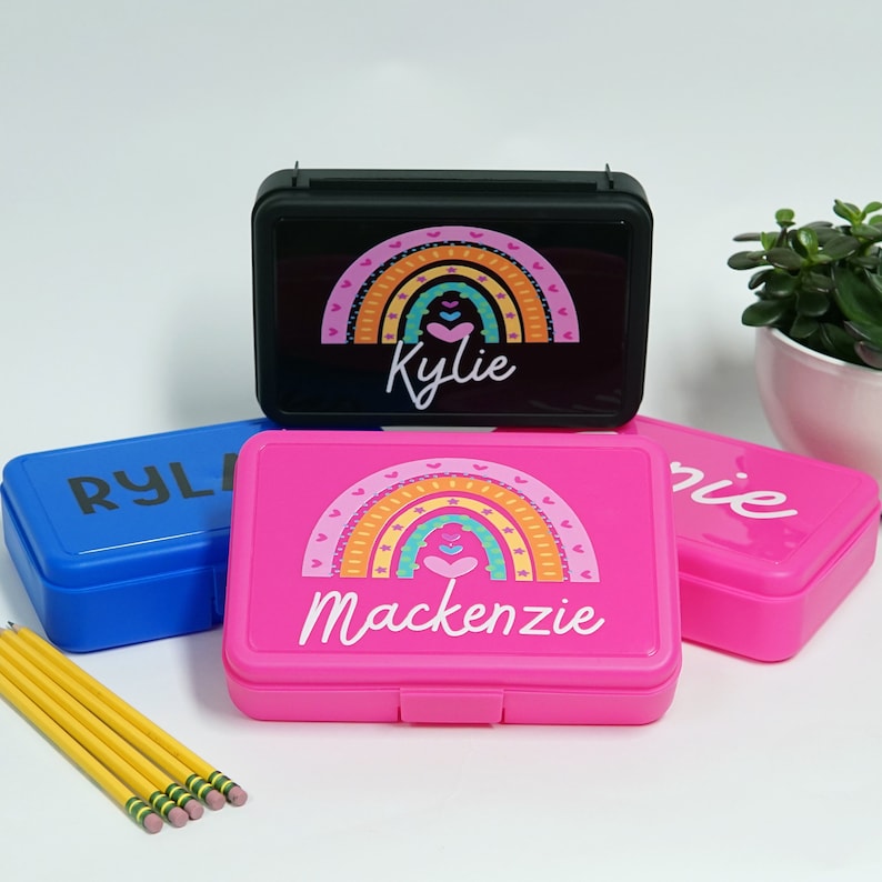 May include: Three colorful plastic pencil boxes with rainbow designs and personalized names. The boxes are stacked on top of each other. The top box is black with the name "Kylie" on it. The middle box is pink with the name "Mackenzie" on it. The bottom box is blue with the name "Ryle" on it.