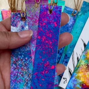 May include: A collection of rectangular paper bookmarks featuring vibrant, multi-coloured abstract paint splatters and patterns. Each bookmark has a hole punched at the top with a thin, light brown twine string attached for hanging or marking pages.