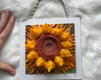 Puede incluir: Una pequeña pintura enmarcada de un girasol con un centro rojo y pétalos amarillos. La pintura está hecha en un estilo texturizado, con la pintura aplicada en capas gruesas. La pintura está colgada en una pared blanca.