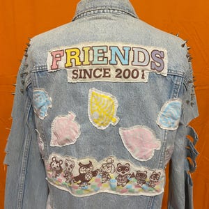 May include: A light blue denim jacket with patches and studs. The patches feature a leaf design, a pink and blue design, and a group of animal characters. The jacket has the text "FRIENDS SINCE 2001" on the back.