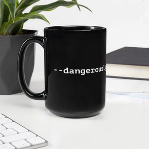 May include: A black ceramic mug with a large handle. The mug features the white text "--dangerously" on its side. The mug is sitting on a white surface, with a plant and a keyboard in the background.