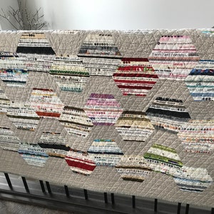 May include: A quilt with a beige background and a pattern of colourful hexagonal shapes. The shapes are made of fabric scraps and have a variety of patterns, including stripes, polka dots, and floral prints. The quilt is quilted with a diamond pattern. The text "Farm Yard Friends" is visible on one of the hexagonal shapes.